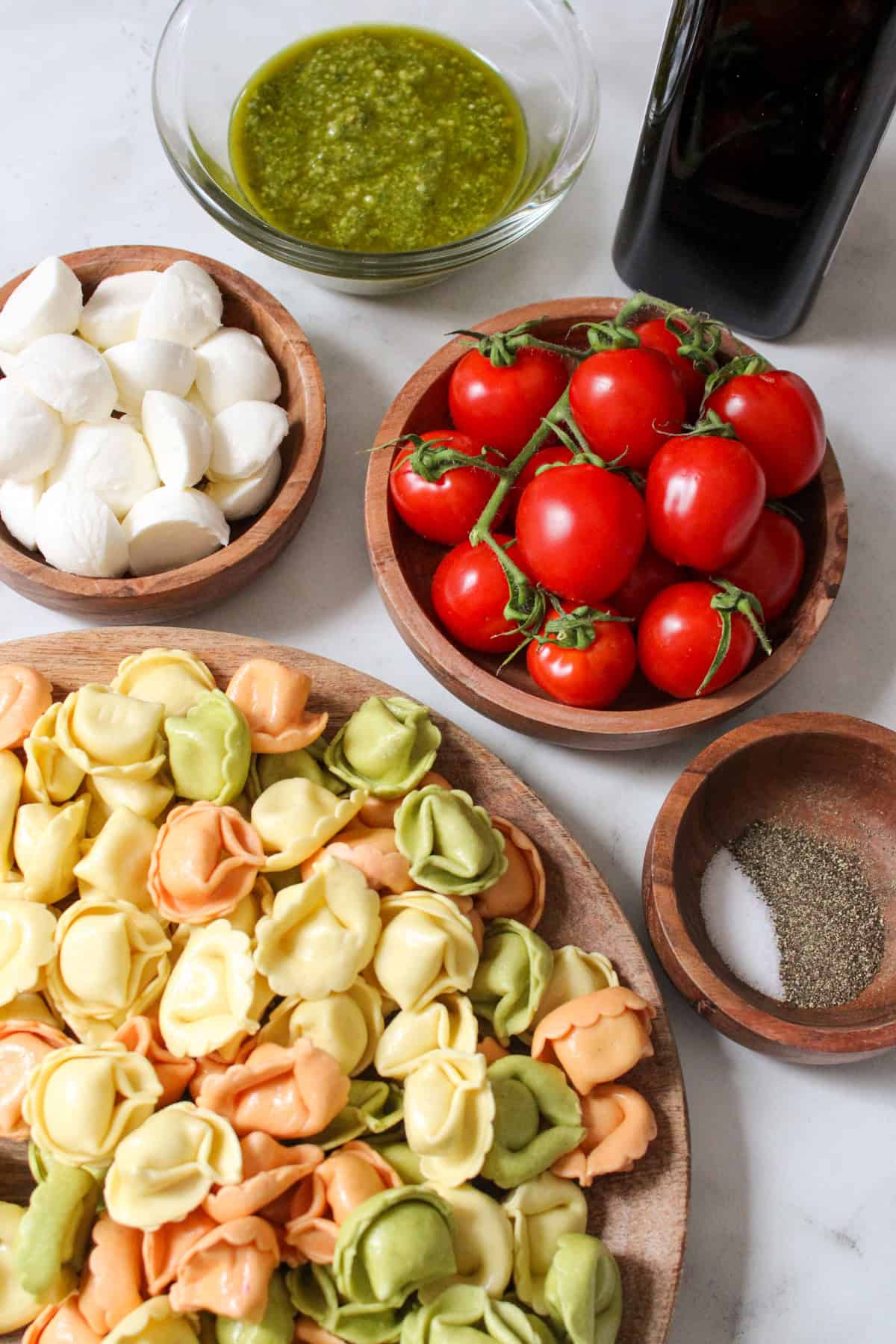 ingredients for caprese tortellini salad in small wooden and glass bowls on a marble counter top