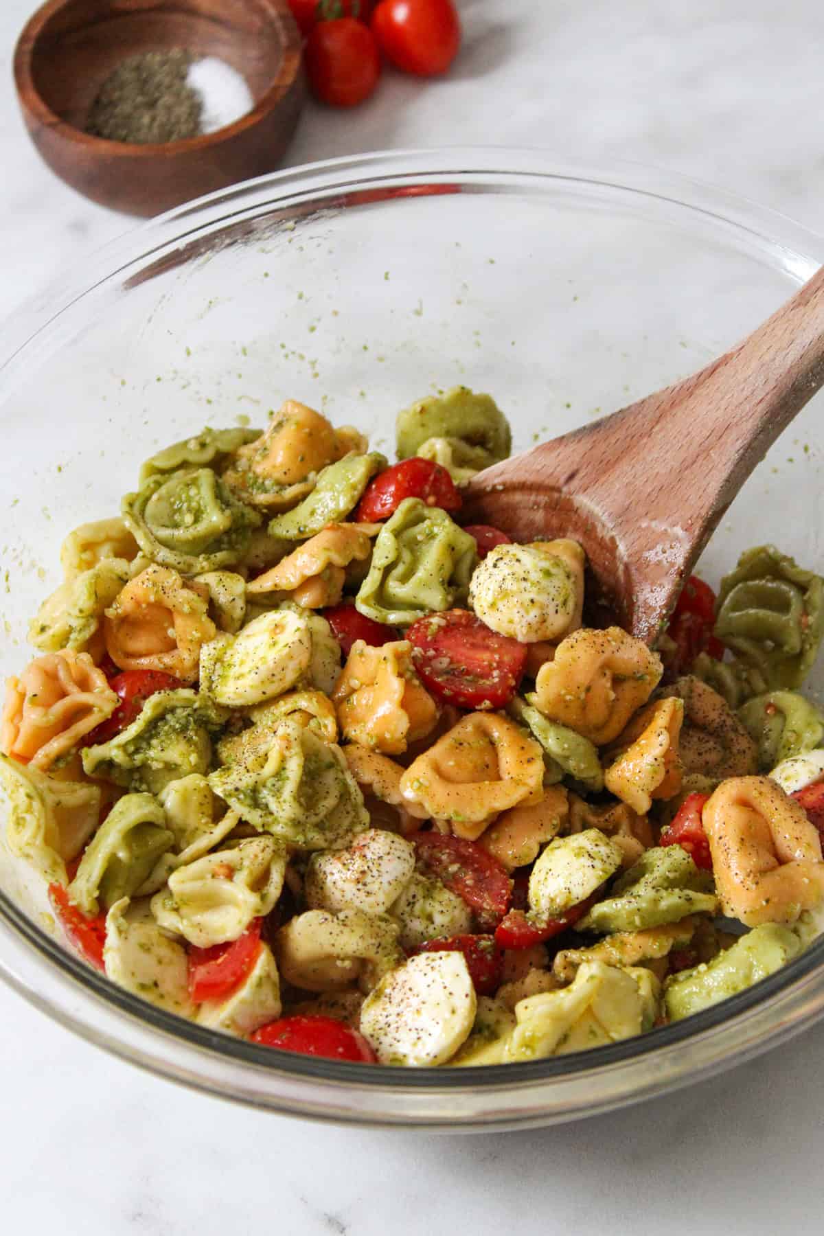 Caprese Tortellini Salad dressed in pesto in a glass bowl with a wooden spoon