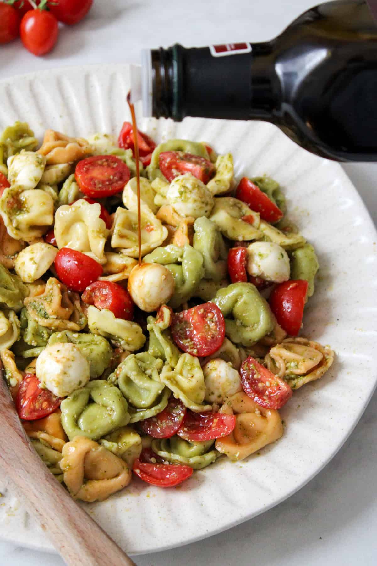 caprese tortellini salad with pesto on a white plate with a wooden spoon, being drizzled with balsamic vinegar