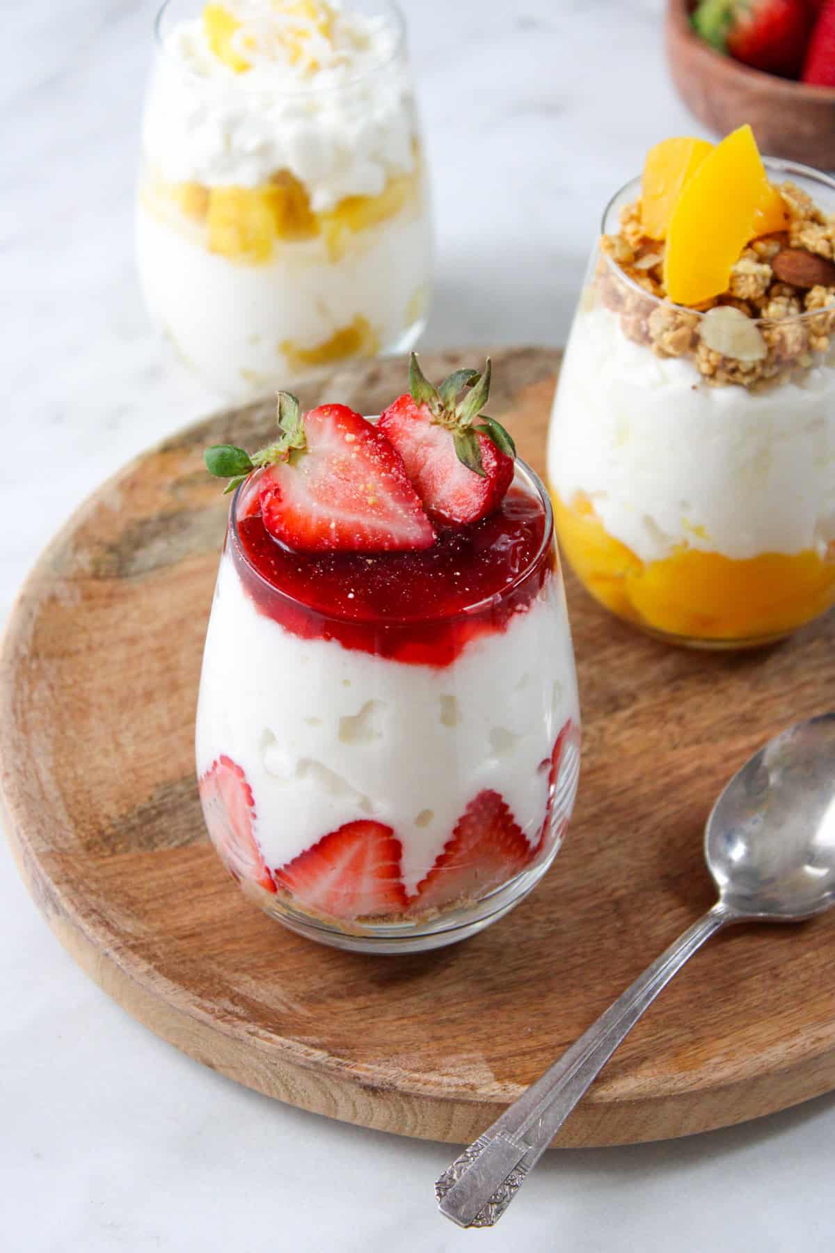 three variations of cottage cheese parfaits with fruit on a wooden platter and marble countertop with a metal spoon