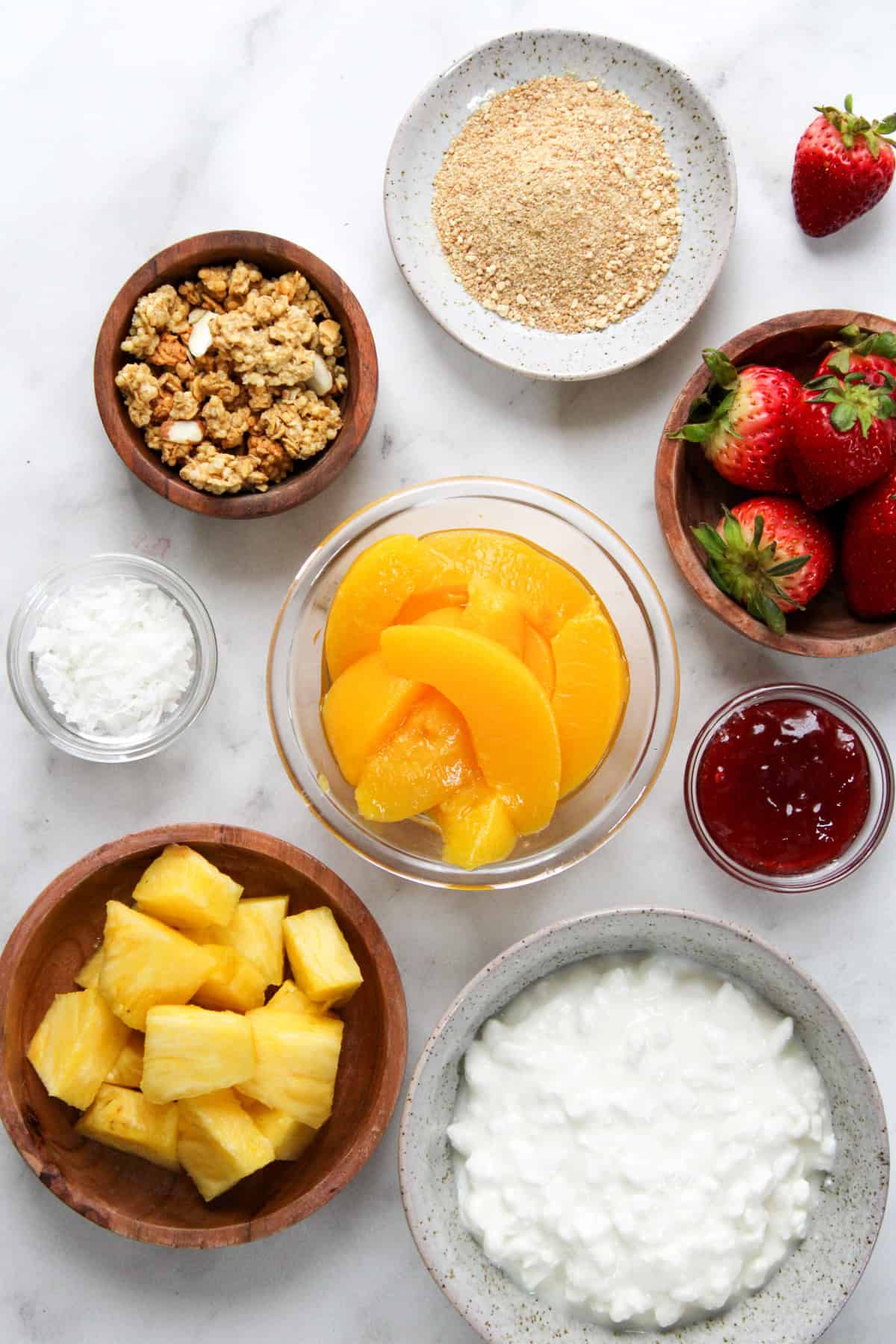 parfait ingredients in small glass and wooden bowls laying on a white marble counter top