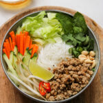 bowl of white noodles, fresh vegetables and herbs on a wooden plate with a small glass bowl of vinaigrette in the background