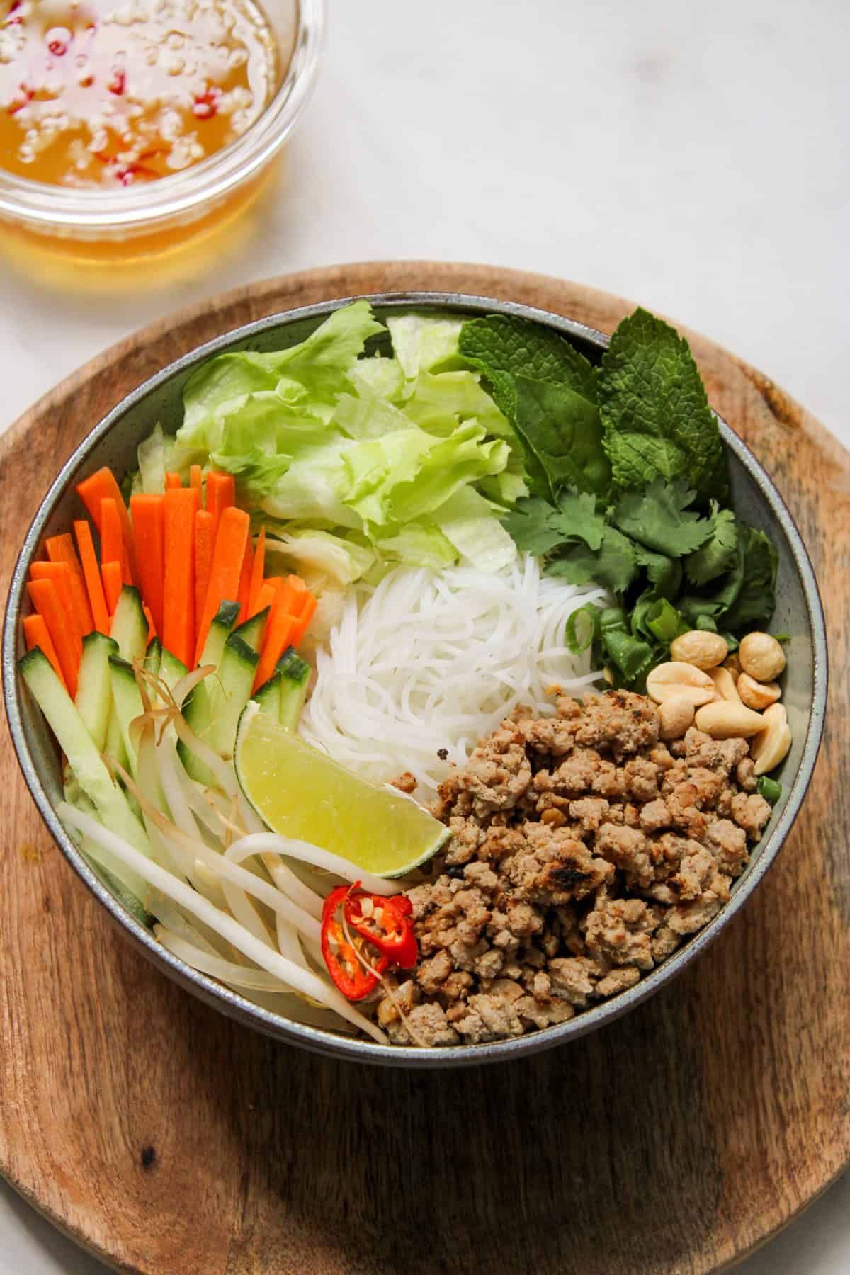 bowl of white noodles, fresh vegetables and herbs on a wooden plate with a small glass bowl of vinaigrette in the background