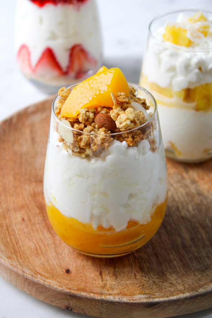 cottage cheese parfait with peaches on the bottom of the glass, topped with granola, almonds, honey and peach, sitting on a wooden platter