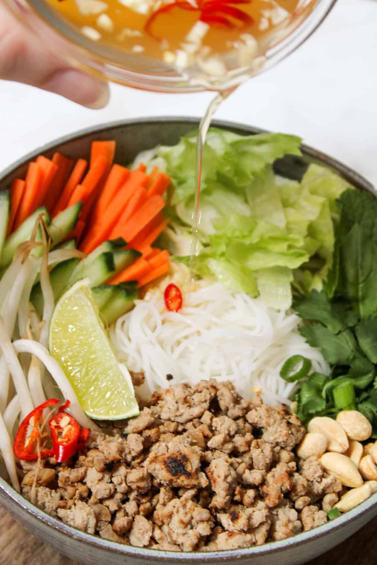 pouring fish sauce vinaigrette with pieces of red chili and garlic from a glass bowl over a ground pork vermicelli bowl