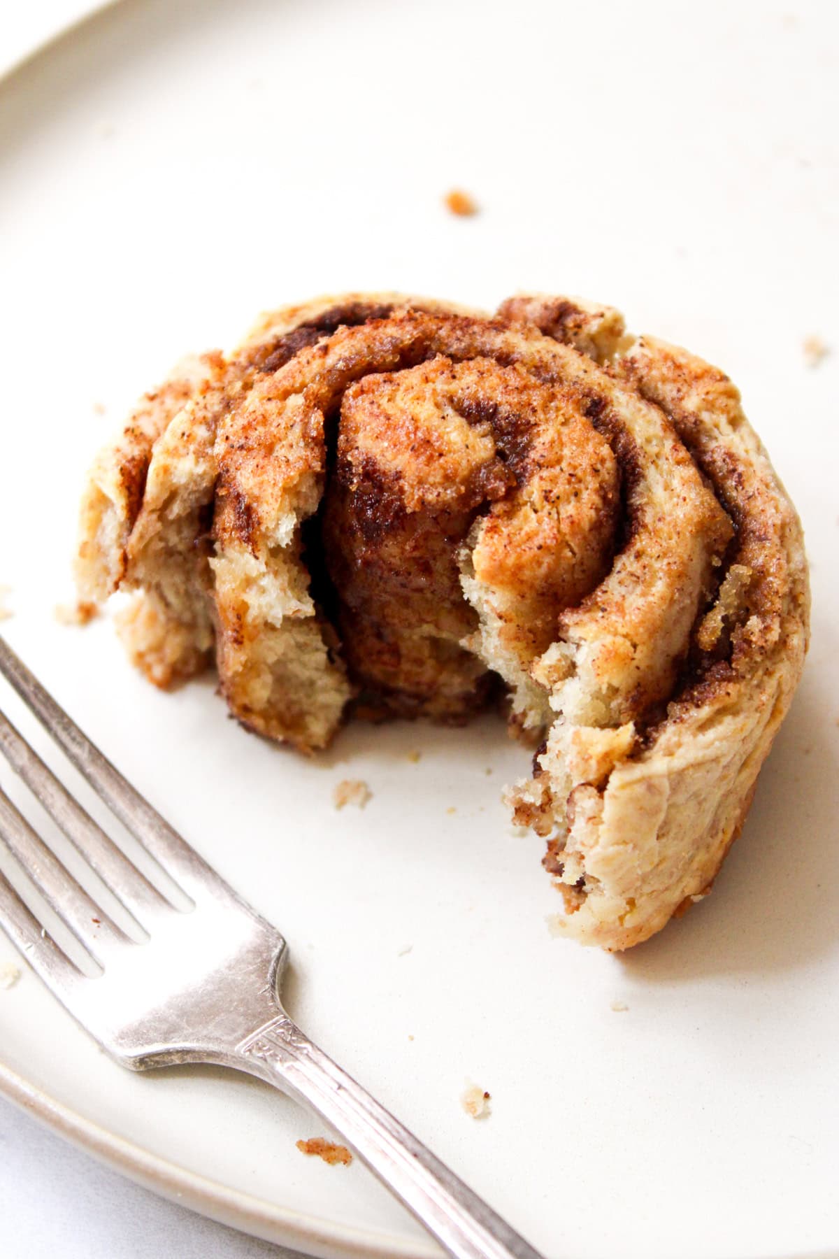 whole wheat cinnamon roll on a beige plate laying next to a fork; the first layers of the cinnamon roll are eaten away to reveal the dark cinnamon center of the roll
