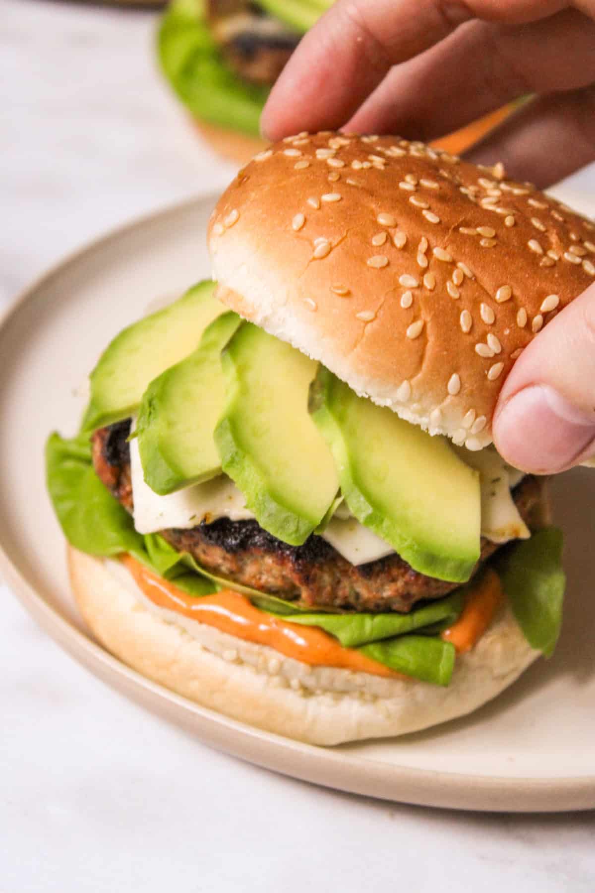 hand placing a burger bun top onto a burger topped with sliced avocado
