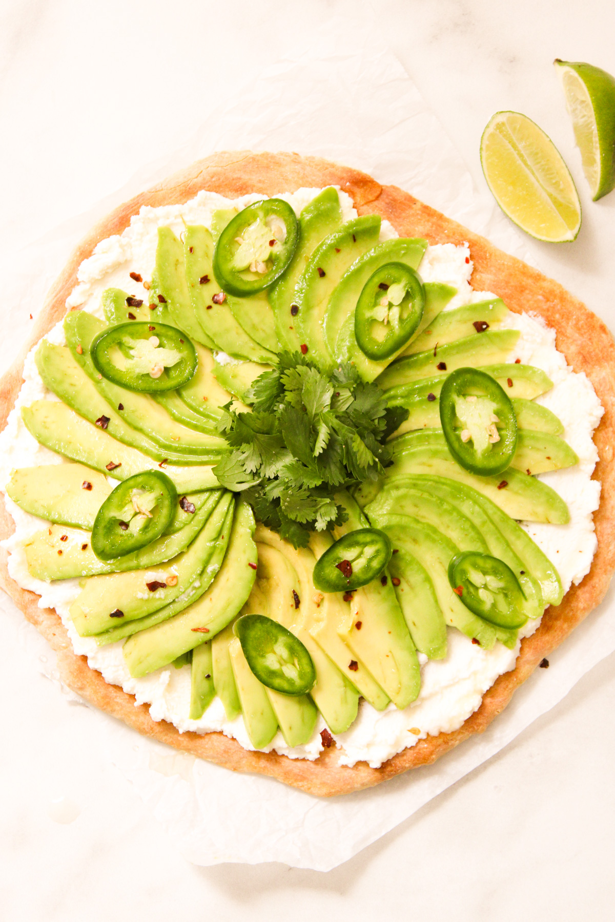 overhead shot of avocado pizza on a piece of parchment laying on a counter beside two wedges of lime