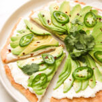 pizza topped with ricotta, fanned avocado, jalapeno and a bundle of cilantro on a white plate