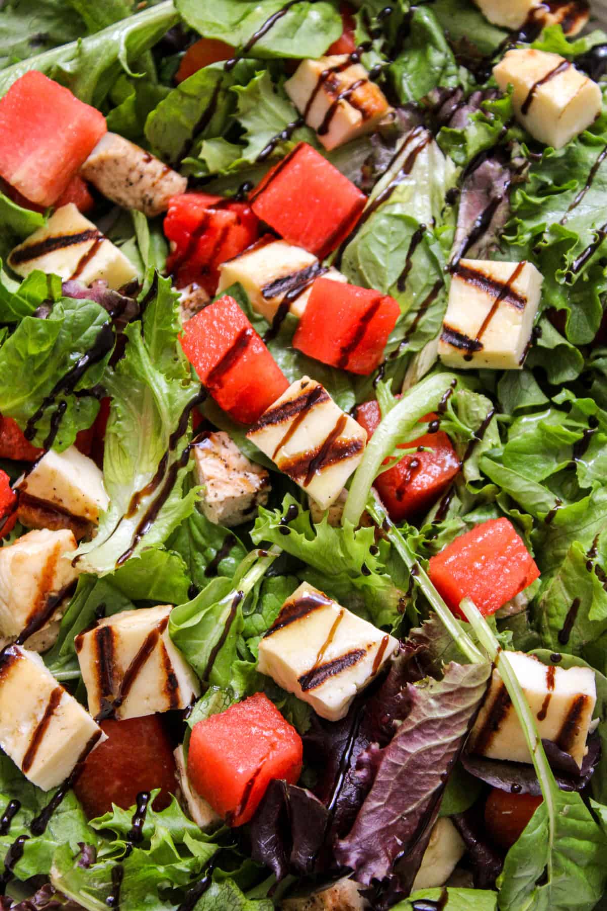 close-up of lettuce topped with cubes of watermelon, chicken and halloumi, drizzled with balsamic glaze