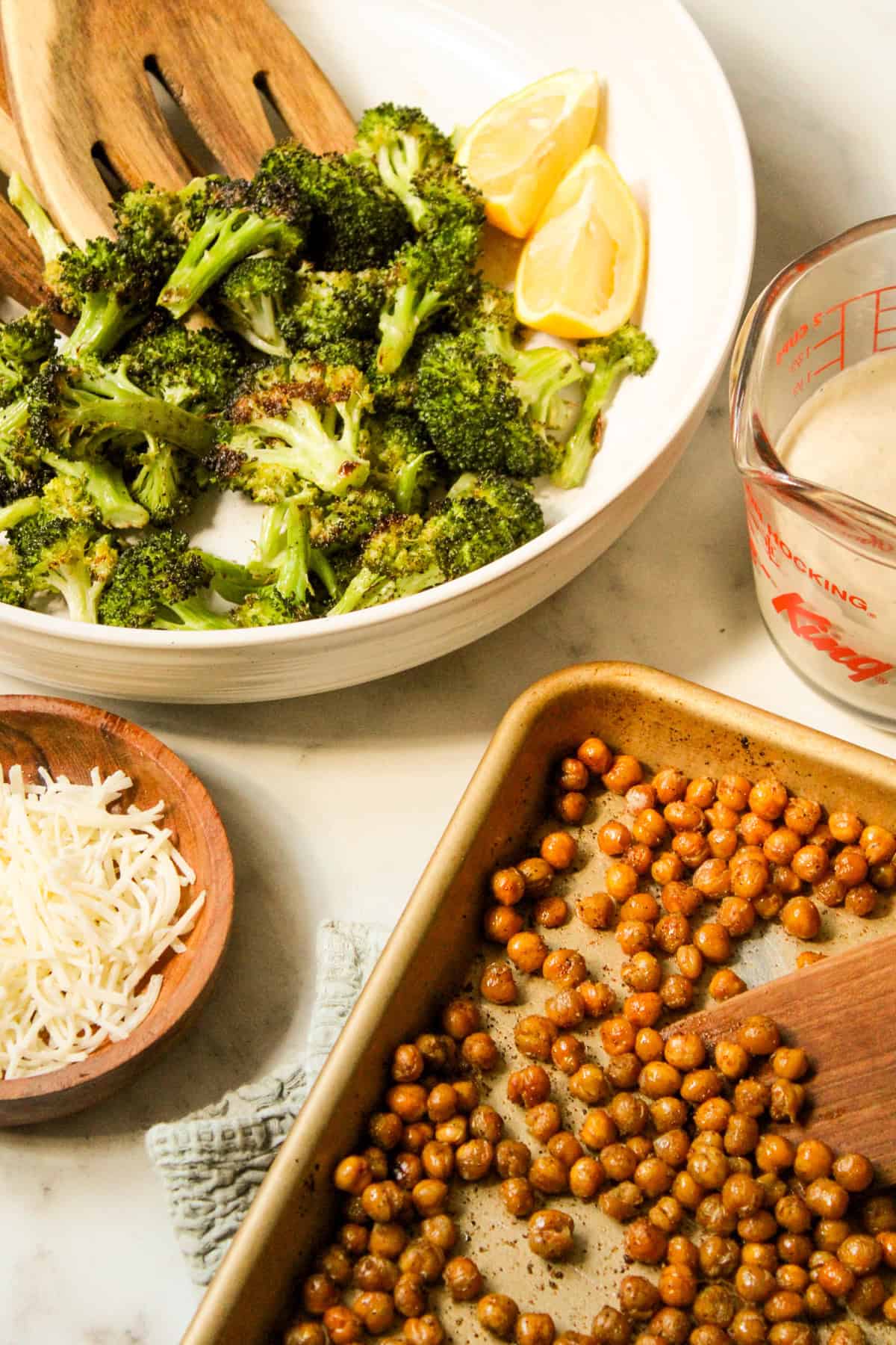 a ceramic bowl with roasted broccoli and lemon wedges alongside a sheet pan of crispy chickpea croutons, parmesan and tahini caesar dressing in a glass measuring glass